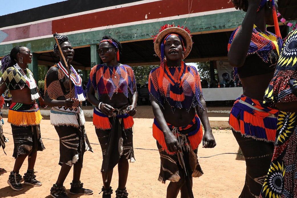 Group of people wearing colorful traditional attire with beadwork and skirts, performing a cultural dance outdoors in front of a painted structure.